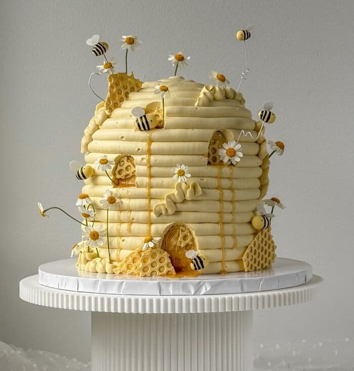 A custom cake sculpted into the shape of a domed beehive using thick horizontal bands of yellow frosting.  The cake is decorated with handcrafted yellow and black bees, some attached directly and others flying on thin wires.  White daisy flowers with yellow centers, textured honeycomb pieces, and realistic honey drips complete the design.