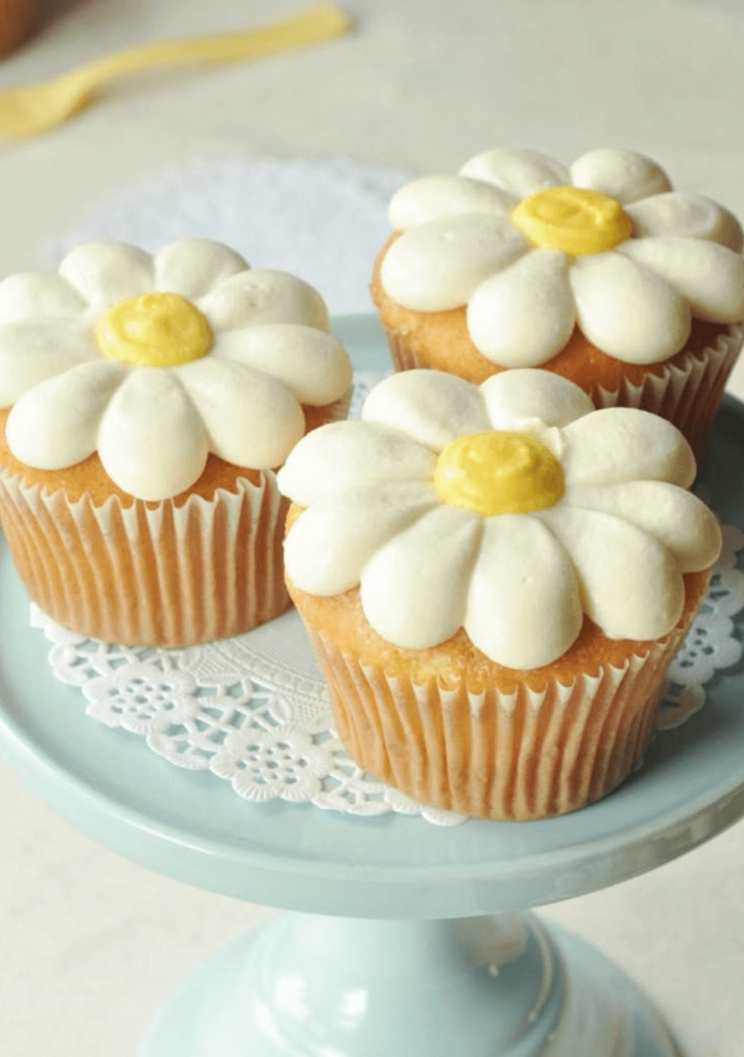 A close-up of a freshly baked cupcake in a white paper liner. The top is generously frosted with a tall swirl of light-colored buttercream, drizzled with thick, golden butterscotch sauce, and heavily sprinkled with crushed, crunchy praline bits. A fluffy butterscotch cupcake topped with swirled frosting, caramel drizzle, and praline crunch.