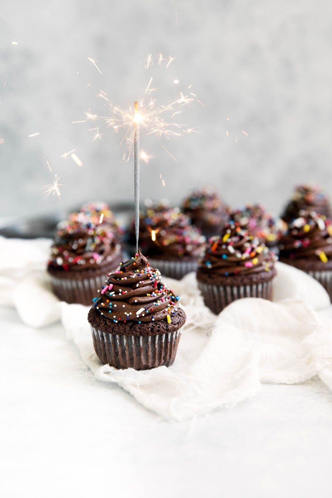 A close-up view of a freshly baked chocolate cupcake sitting in a dark paper liner. The top of the cake is generously frosted with a tall, smooth swirl of dense, dark chocolate truffle ganache. The frosting is lightly dusted with fine cocoa powder and finished with delicate chocolate shavings resting on the peak. Handcrafted chocolate truffle cupcake by PlateStory in Chennai, featuring a moist cocoa sponge topped with dense chocolate ganache frosting, cocoa dusting, and chocolate curls.