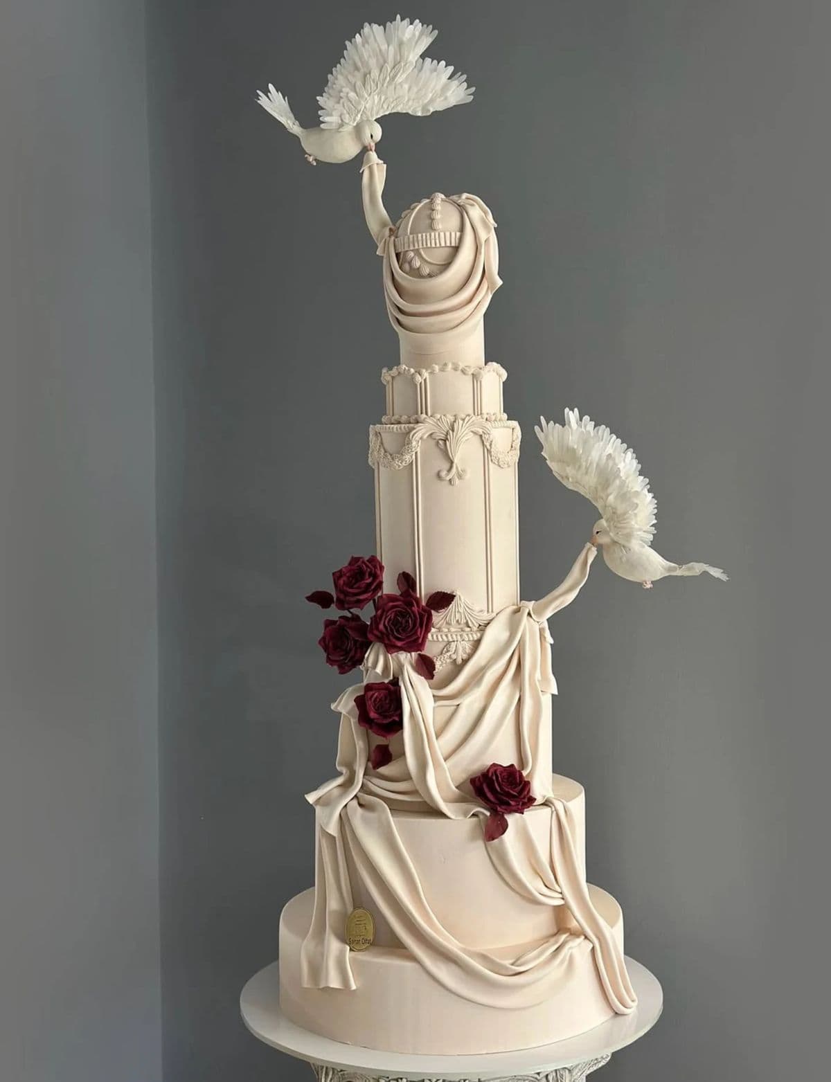 A tall, multi-tiered cream cake featuring classical architectural details, dark red roses, and sculpted white doves holding draped fondant. A towering cream-colored custom cake with multiple tiers. The tiers feature ornate architectural moldings and paneling. Deep red sugar roses cascade down the middle sections, while two sculpted white doves with feathered wings appear to be holding up pieces of realistic, draping fondant fabric that wrap around the cake.