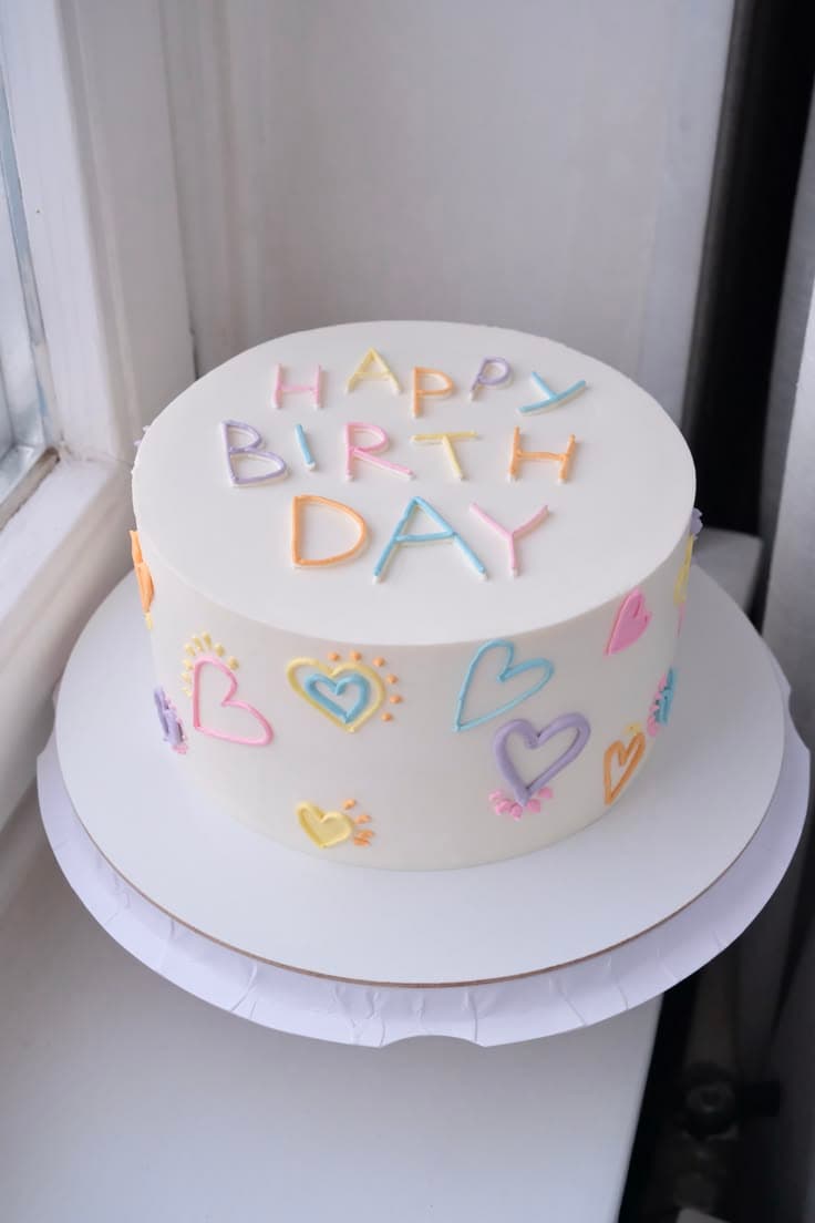 A high-angle view of a round white frosted cake resting on a white cake board by a window. The top features "HAPPY BIRTHDAY" piped in pastel pink, yellow, orange, purple, and light blue letters. The sides are decorated with hand-piped pastel outline hearts with tiny sunburst details in matching colors.