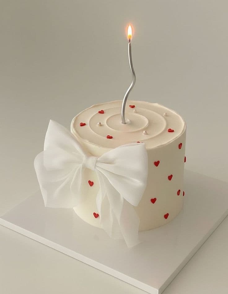 A high-angle view of a round, cream-colored bento cake on a white square board. The cake features a pattern of small red piped hearts and a large white silk fabric bow attached to the front. A single lit, wavy silver candle stands in the center of the spiral-textured top, accented with tiny white pearls. The background is a clean, off-white
