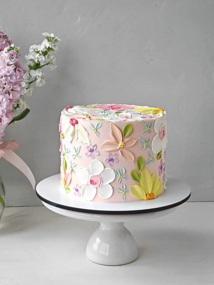 A tall, round birthday cake frosted in soft blush pink buttercream, resting on a white pedestal stand. The cake is decorated with thick, 3D palette-knife-sculpted flowers and leaves in shades of white, peach, yellow, purple, and blue-green, giving it an oil painting texture. Pink flowers in a glass vase sit blurred to the left against a grey background.