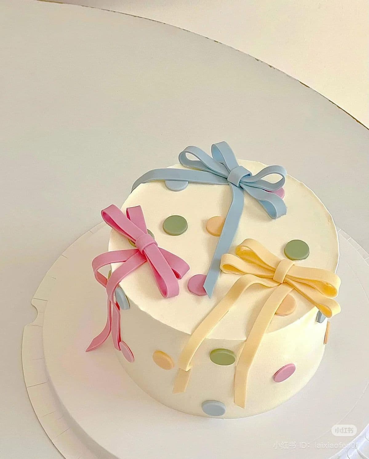 A high-angle view of a round cake frosted in smooth ivory buttercream, resting on a white board. The cake is decorated with three 3D pastel fondant bows in pink, blue, and light yellow, along with flat pastel polka dots in green, yellow, pink, and blue scattered across the top and sides.