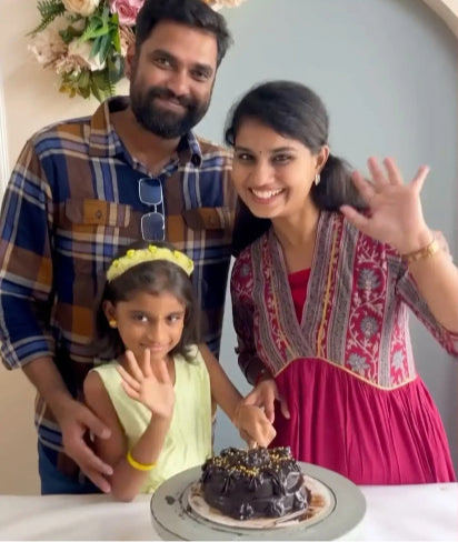 Family of three with a chocolate cake on a table, smiling and waving.